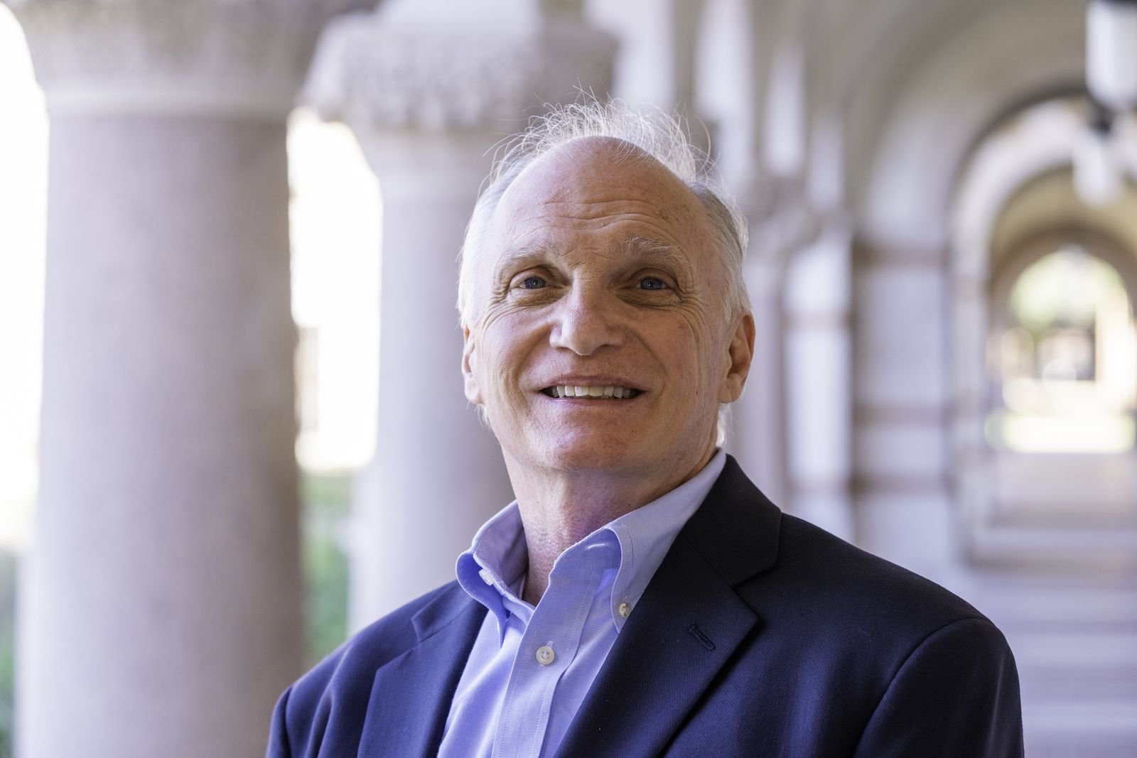 Skip York in a stone colonnade, smiling, with arches receding into daylight behind him